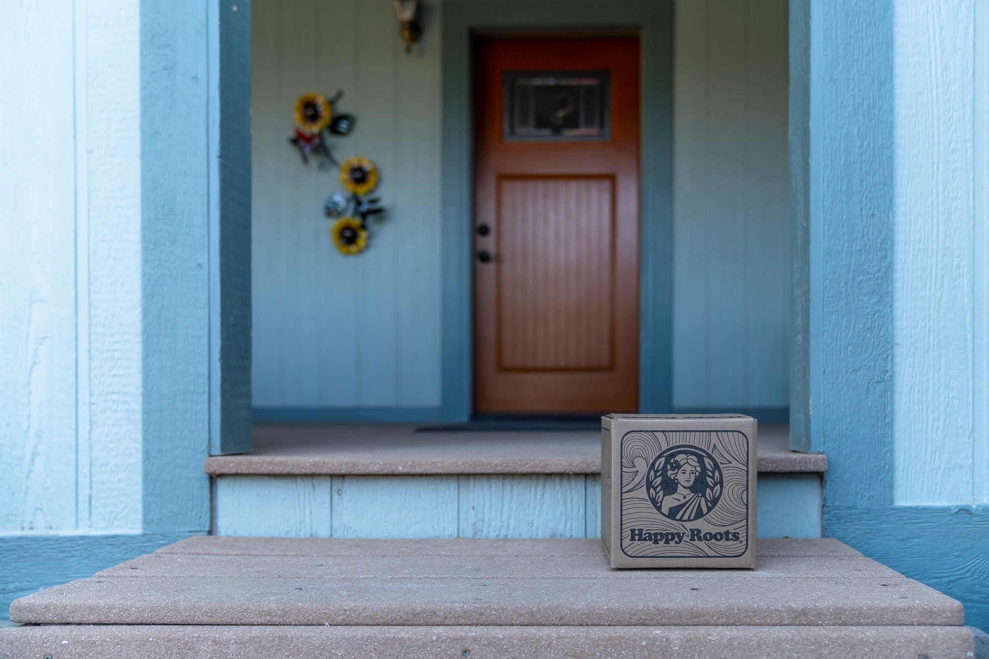 A Happy Roots delivery box placed on a front porch with a wooden door and sunflower wall decorations in the background.