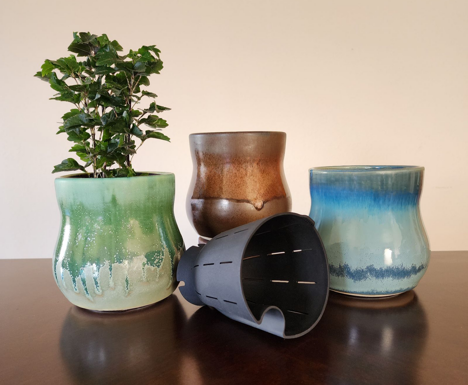 Three ceramic Happy Roots Plant Pots are displayed on a dark wooden surface. The 'Evergreen' pot on the left holds a leafy green plant, featuring a glossy glaze transitioning into a matte finish with flowing drips of light green and cream. The 'Copper Ore' pot in the center has a rich brown glaze with metallic highlights and a smooth texture. The 'Frozen Creek' pot on the right features a glossy glaze blending from icy blue at the top to a darker, textured base. A flexible removable liner in front.
