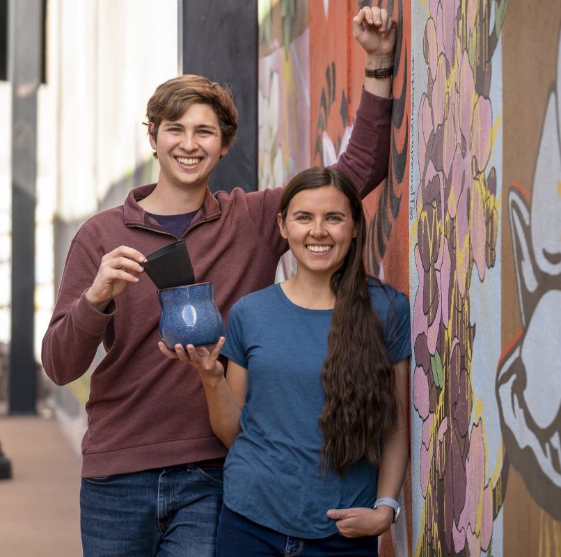 Marc Fiot and Rikki Cook standing outdoors against a colorful mural wall, holding a Happy Roots Plant Pot.