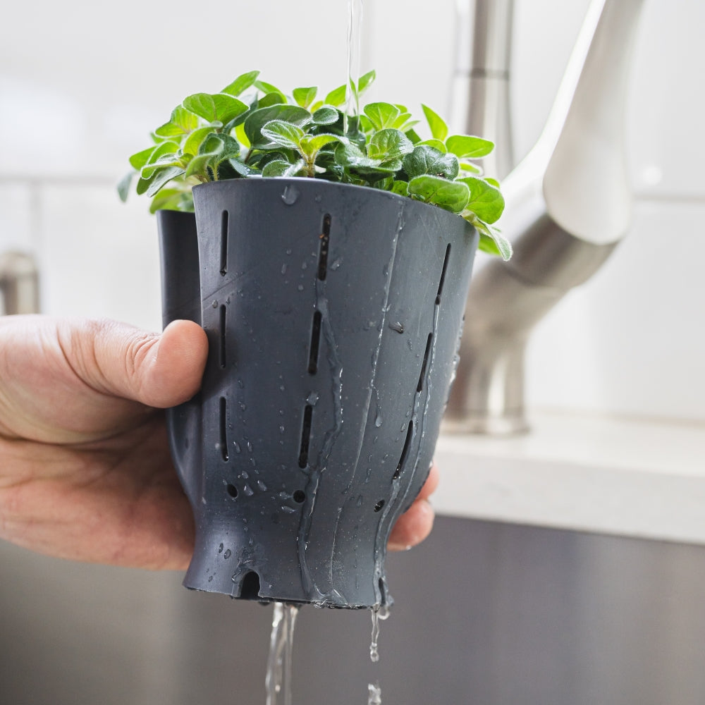 Hand holding a black Happy Roots plant pot liner under a faucet with water flowing through its drainage slits, showcasing its innovative and functional design.