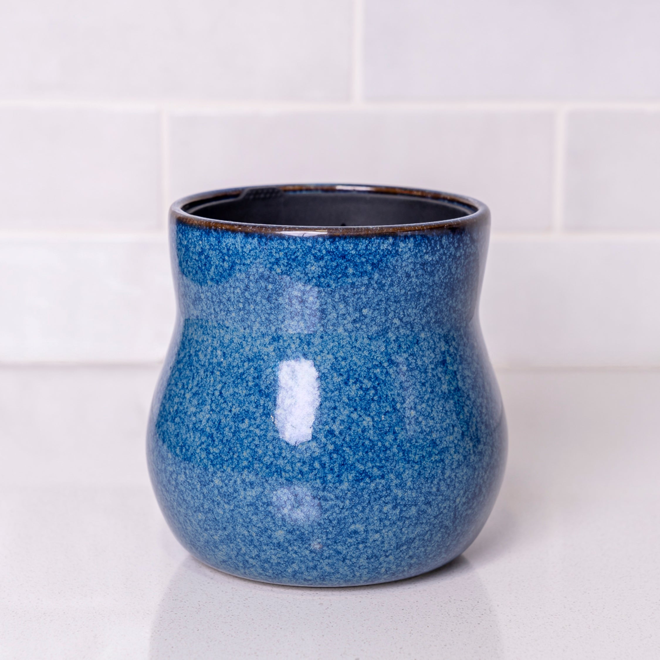 Mottled river blue curvy Happy Roots plant pot on a speckled white counter in front of white kitchen tiles.