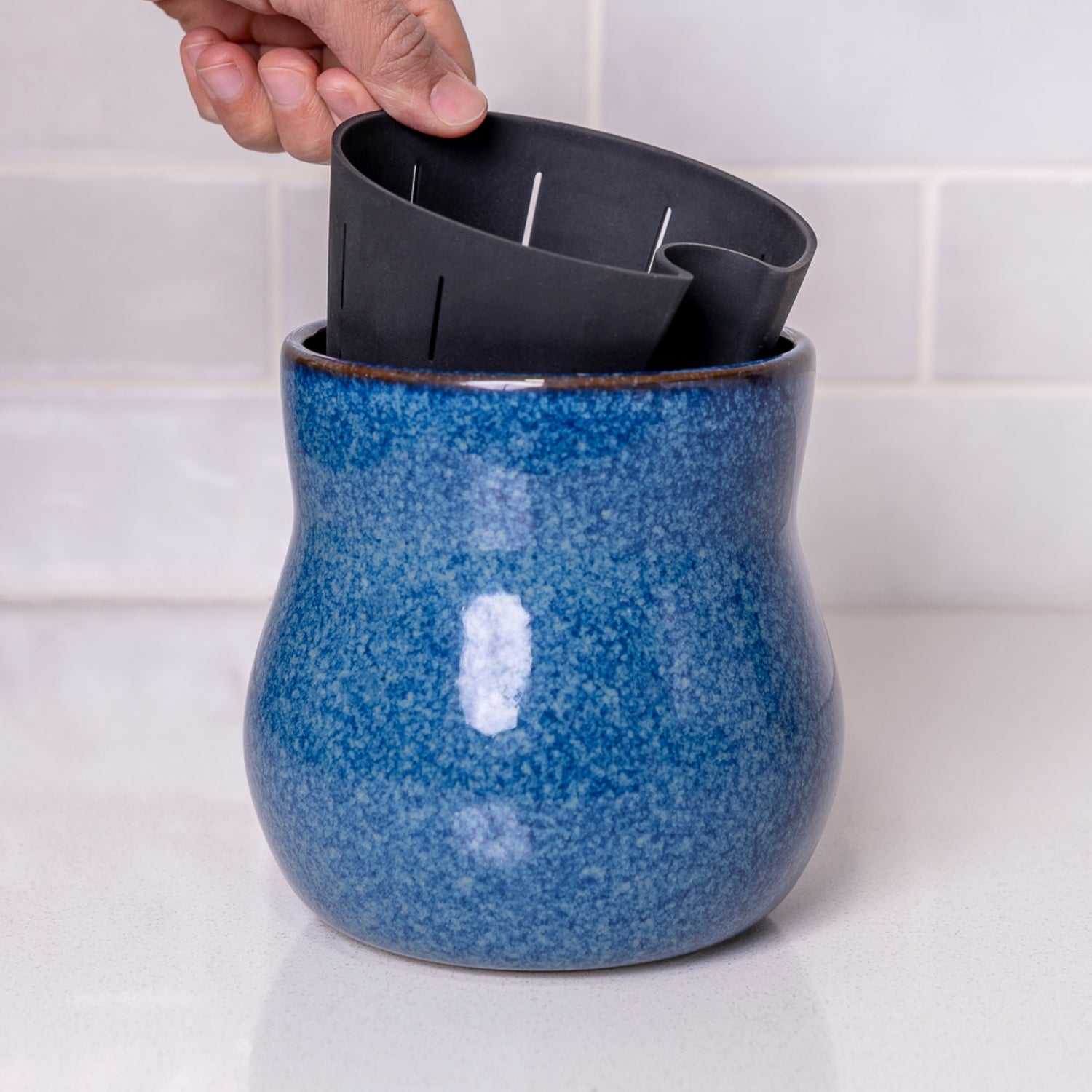 Hand lifting liner from a curvy mottled blue Happy Roots plant pot on a white counter in front of white tiles.