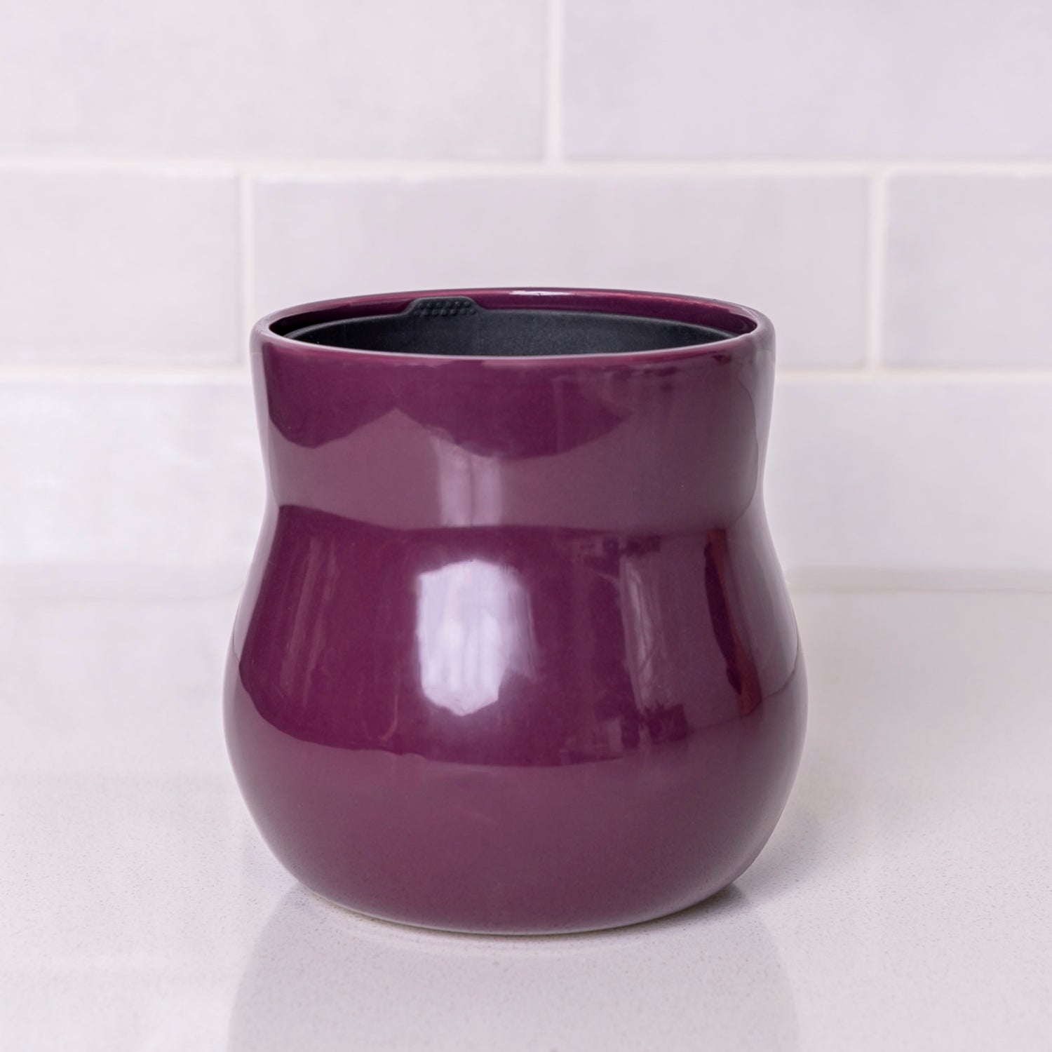 A curvy purple violet Happy Roots Plant Pot on a white counter in front of white kitchen tiles.